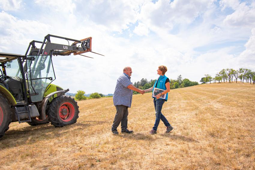 Naturtalent Stefanie Schwarz begrüßt einen Landwirt per Handschlag auf einer Anhöhe, neben ihnen befindet sich ein Traktor.