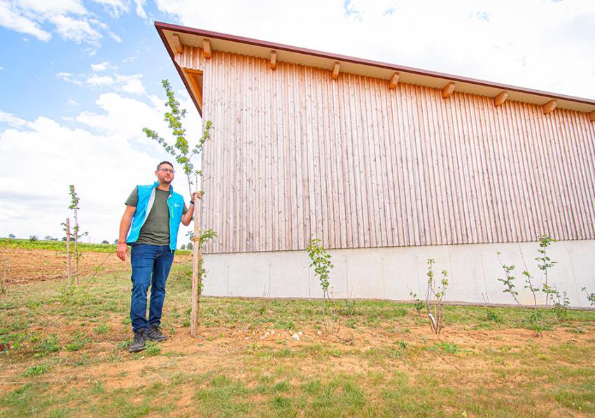Naturtalent Christof Simon steht vor einer neugebauten Maschinenhalle, um ihn herum befinden sich neugepflanzte Bäume als Ausgleichsmaßnahme für den Neubau.