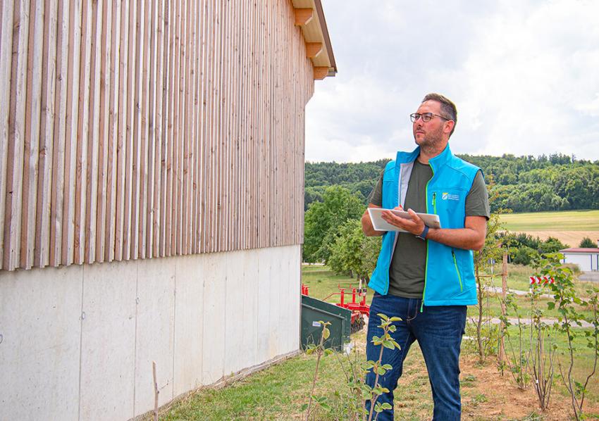 Naturtalent Christof Simon steht neben einer neu erbauten landwirtschaftlichen Gerätehalle und macht Notizen.