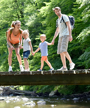 Das Bild zeigt eine Familie, die auf einer Holzbrücke über einen Bach geht