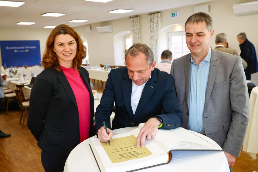 Umweltminister Thorsten Glauber steht am Stehtisch und schreibt in das Gästebuch, links davon Tanja Schweiger und rechts steht Josef Schindler