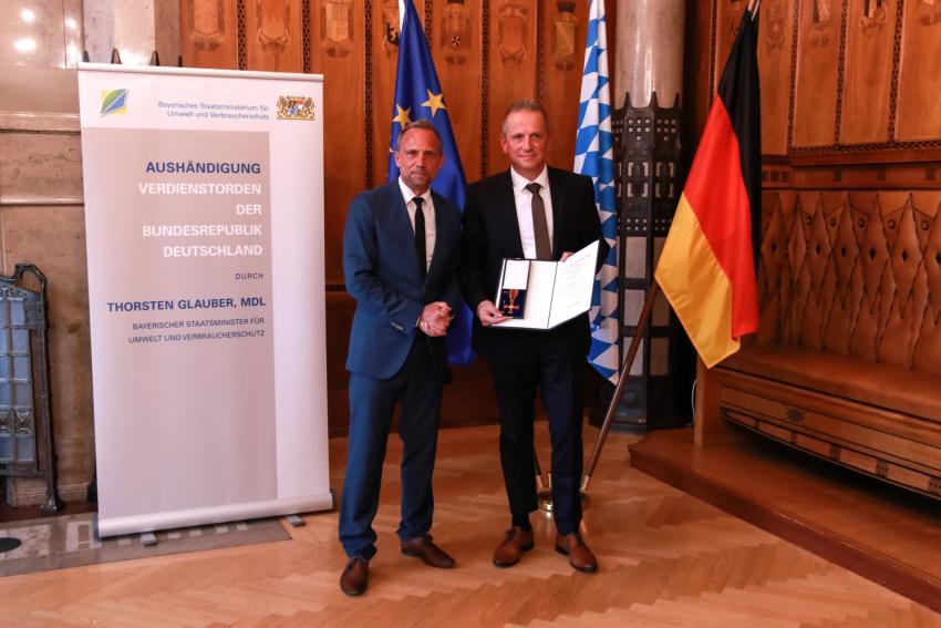 Das Foto zeigt Umweltminister Thorsten Glauber (li) zusammen mit dem neuen Ordensträger Wolfgang Heß, er zeigt die Verleihungsurkunden und das Bundesverdienstkreuz in die Kamera. ©Foto Benjamin Böhm