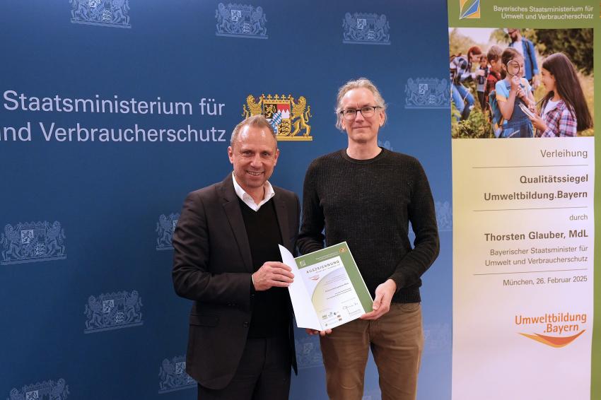 Umweltminister Thorsten Glauber mit einem Vertreter des Naturerlebniszentrum Rhön in Hammelburg, zusammen zeigen sie die Auszeichnungsurkunde.