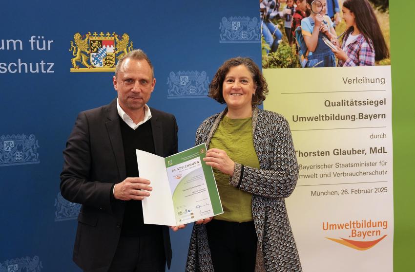 Umweltminister Thorsten Glauber mit einer Vertreterin vom Transition Town Augsburg e.V., zusammen zeigen sie die Auszeichnungsurkunde.