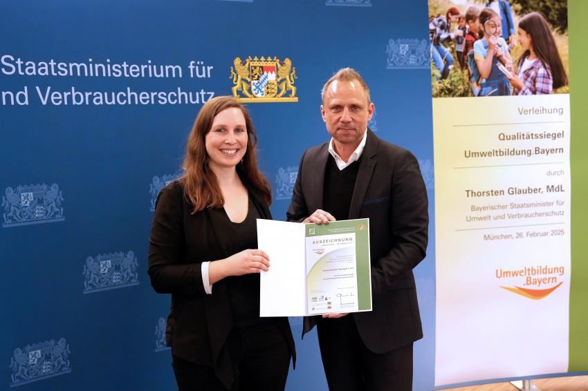 Umweltminister Thorsten Glauber mit einer Vertreterin von der Umweltstation Kitzinger Land aus Marktsteft, zusammen zeigen sie die Auszeichnungsurkunde.