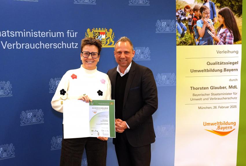 Umweltminister Thorsten Glauber mit einer Vertreterin der Almschule im Werksviertel Mitte in München, zusammen zeigen sie die Auszeichnungsurkunde.
