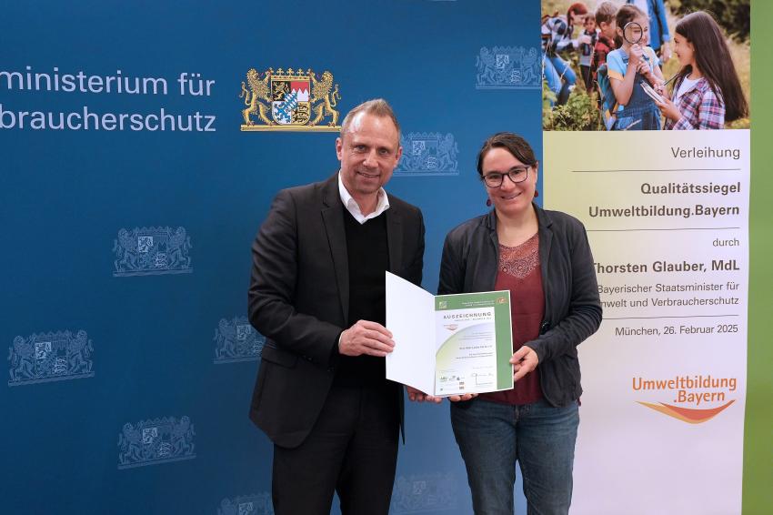 Umweltminister Thorsten Glauber mit einer Vertreterin vom Eine Welt Laden Fürth e.V., zusammen zeigen sie die Auszeichnungsurkunde.