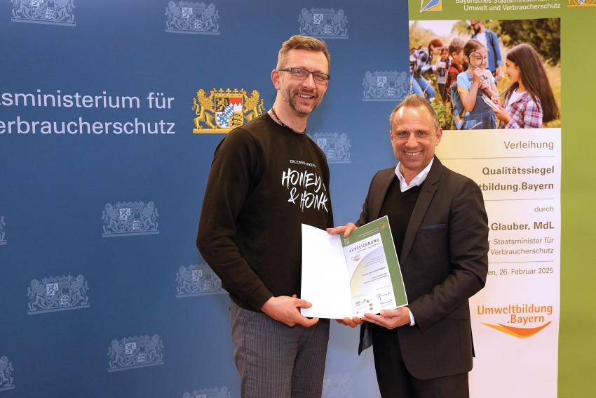 Umweltminister Thorsten Glauber mit einem Vertreter der Erlebnis-Imkerei Honey&Honk in Nürnberg, zusammen zeigen sie die Auszeichnungsurkunde.