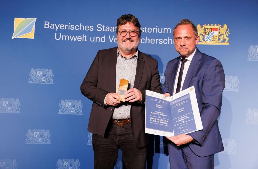 Umweltminister Thorsten Glauber und ein Vertreter der Gemeinde Fuchstal zeigen zusammen den Klimaschutzpreis und die Urkunde.