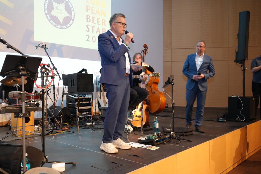 Auf dem Foto zu sehen Umweltminister Thorsten Glauber (re.) und Stefan Stang (Geschäftsführer Private Brauereien Bayern und Deutschland) bei einer Rede und im Hintergrund eine Musikband.