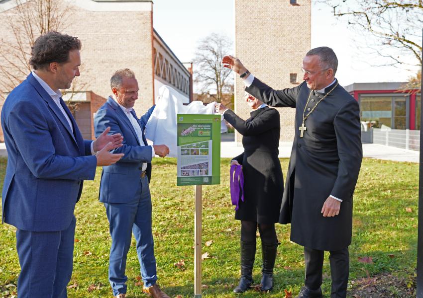 Bürgermeister Dr. Andreas Bukowski, Staatsminister Thorsten Glauber, Pfarrerin Annedore Becker und Landesbischof Christian Kopp enthüllen das neu aufgestellte Blühpakt-Schild auf dem Vorplatz der Jesuskirche in Haar. 