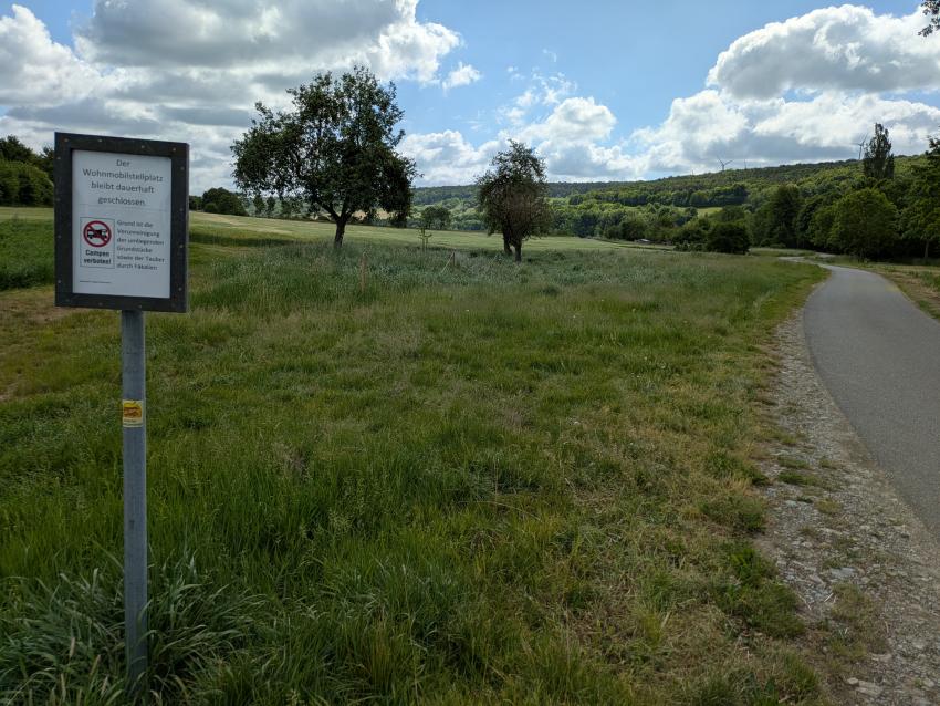 Ansicht der Teilfläche 3 nach 2 Jahren, im Mai 2025 nach der Maßnahmenumsetzung. Um die Wiese mit mehrjährigen Wiesenblumen anzureichern, wurde auf einem Streifen gebietsheimisches Saatgut eingebracht. Der Campingplatz wurde wegen der andauernden Müllproblematik geschlossen. Das Streuobst wurde mit einer Birne erweitert, außerdem blieb das  Totholz der alten Bäume erhalten und bietet so Lebensraum für zahlreiche Insekten und andere Tierarten. 