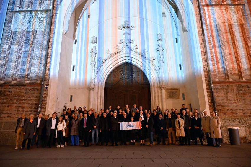 Gruppenfoto mit den Partnerinnen und Partnern der Bayerischen Klima-Allianz vor dem Münchner Liebfrauendom, der zum Start der bayerischen Klimawoche in den Farben der bayerischen Klimastreifen leuchtet.