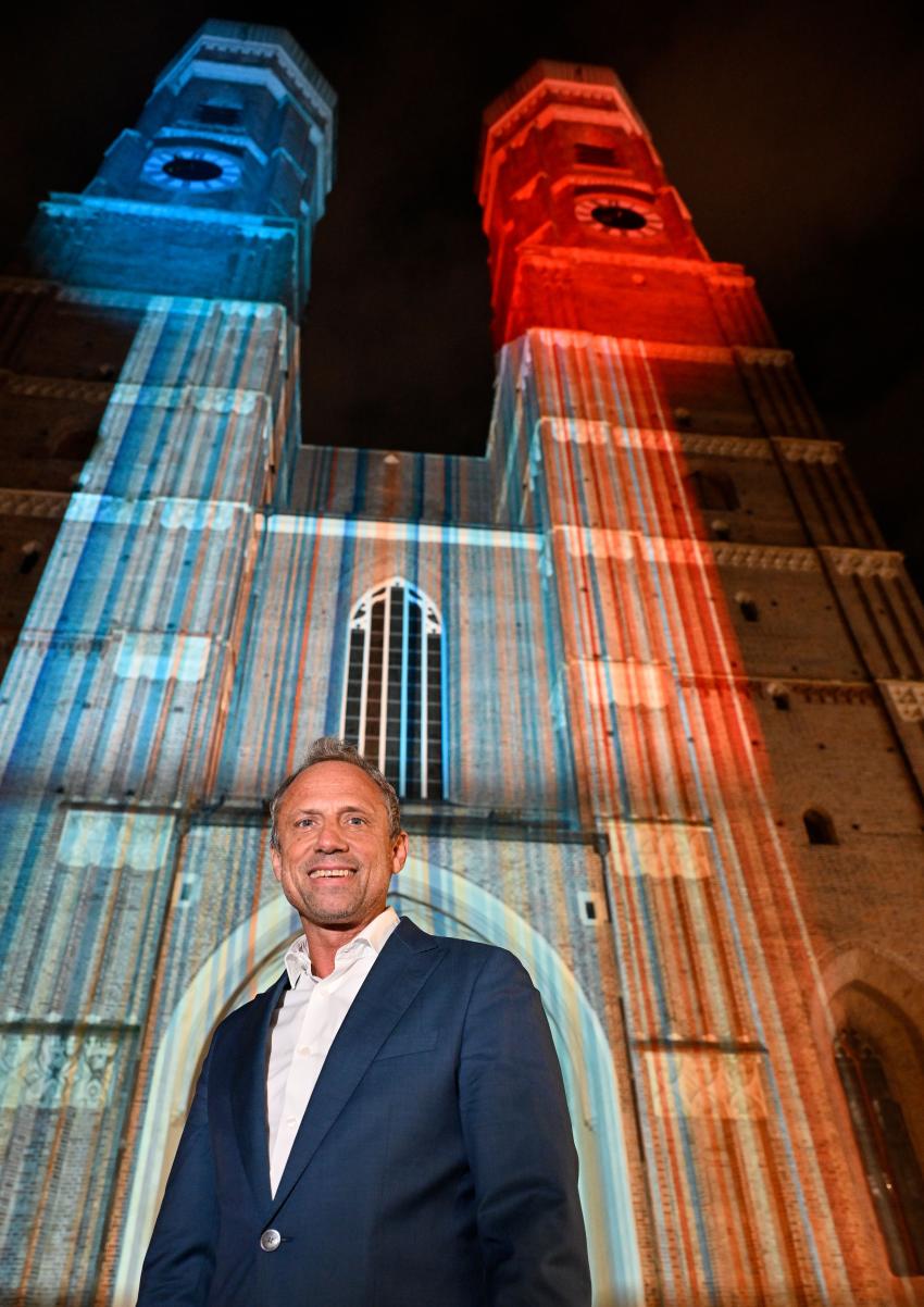 Umweltminister Thorsten Glauber vor dem Münchner Liebfrauendom, der zum Start der bayerischen Klimawoche in den Farben der bayerischen Klimastreifen leuchtet.