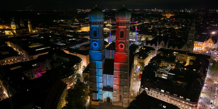 Drohnenaufnahme des Münchner Liebfrauendoms, der in den Farben der bayerischen Klimastreifen leuchtet