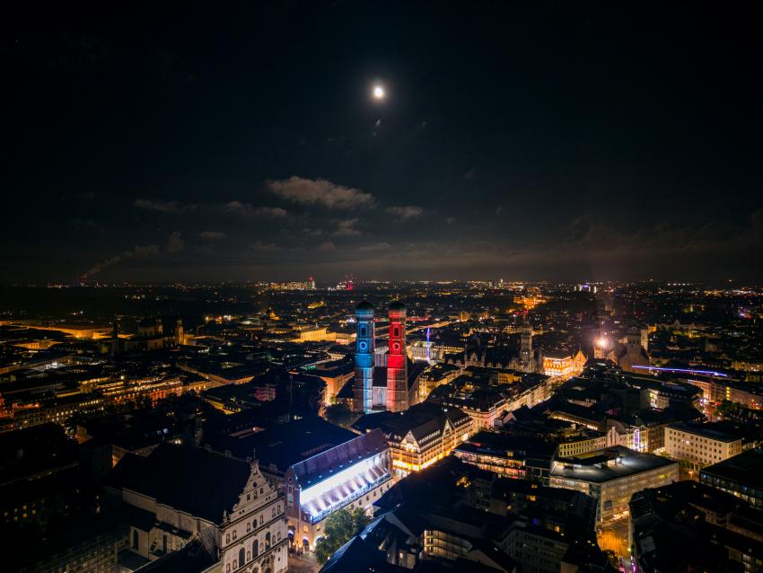 Drohnenaufnahme des Münchner Liebfrauendoms, der in den Farben der bayerischen Klimastreifen leuchtet