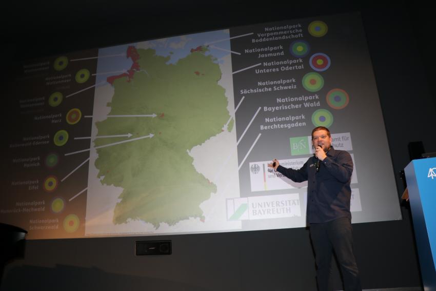 Das Foto zeigt Dr. Benedikt Wiggering auf der Bühne bei seinem Kurzvortrag „Wer fraß am Aas?“ (Deutscher Science Slam Meister 2024 und Mitarbeiter NPV Niedersächsisches Wattenmeer).