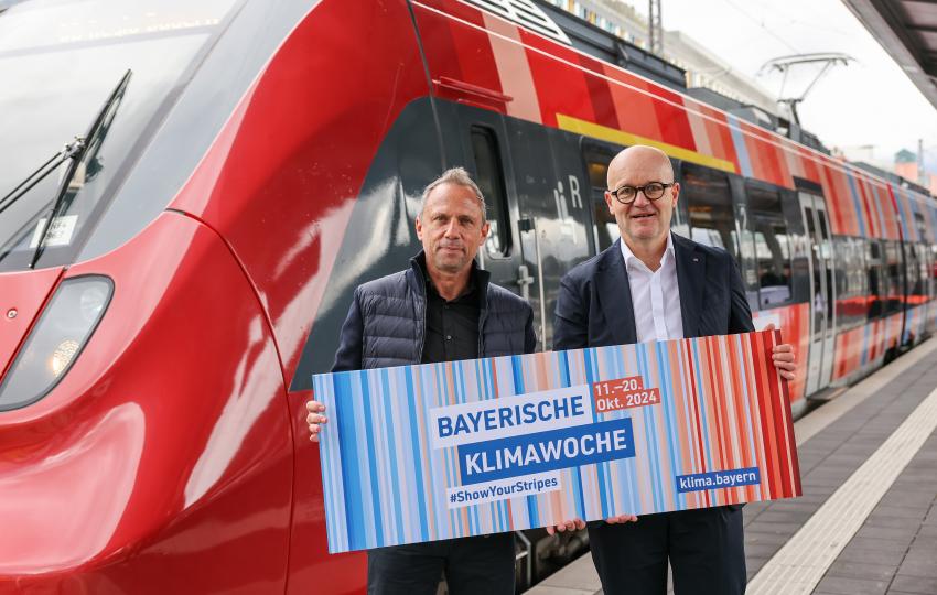 Das Bild zeigt Bayerns Umweltminister Thorsten Glauber zusammen mit Heiko Büttner, DB-Konzernbevollmächtigter für den Freistaat Bayern, vor dem Streifenzug stehen. Sie halten das Banner zur Eröffnung der bayerischen Klimawoche.