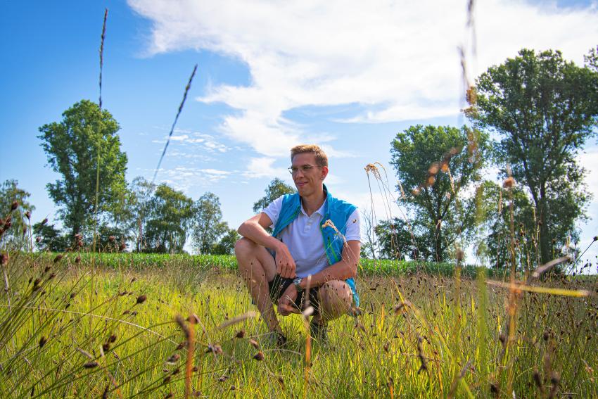 Naturtalent Aaron Hoyer betrachtet in der Hocke die Pflanzen in einer hochgewachsenen Wiese.