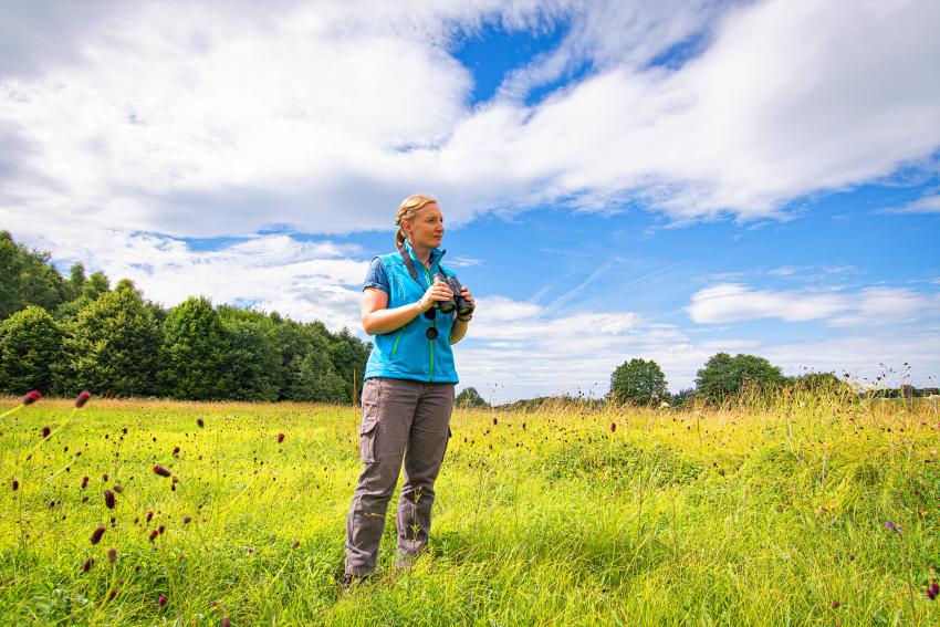 Naturtalent Manuela Kern steht mit einem Fernglas in Ihren Händen inmitten einer hochgewachsenen Wiese.