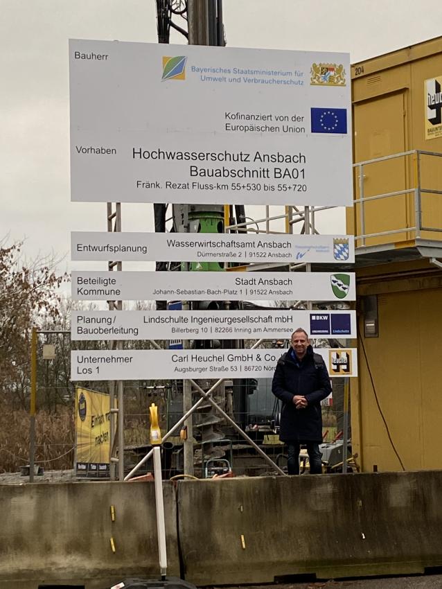 Minister Glauber steht auf der Baustelle vor dem Schild für Ausweisung des Bauvorhabens.
