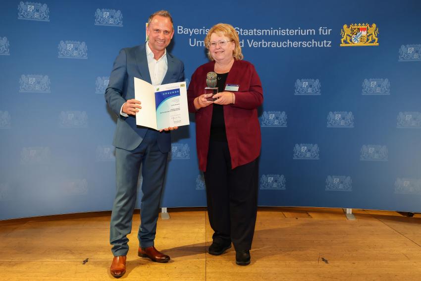 Das Foto zeigt den Bayerischen Umweltminister Thorsten Glauber mit der Tierschutzpreisträgerin Ilona Wojahn, die Auszeichnungsurkunde und den Tierschutzpreis.