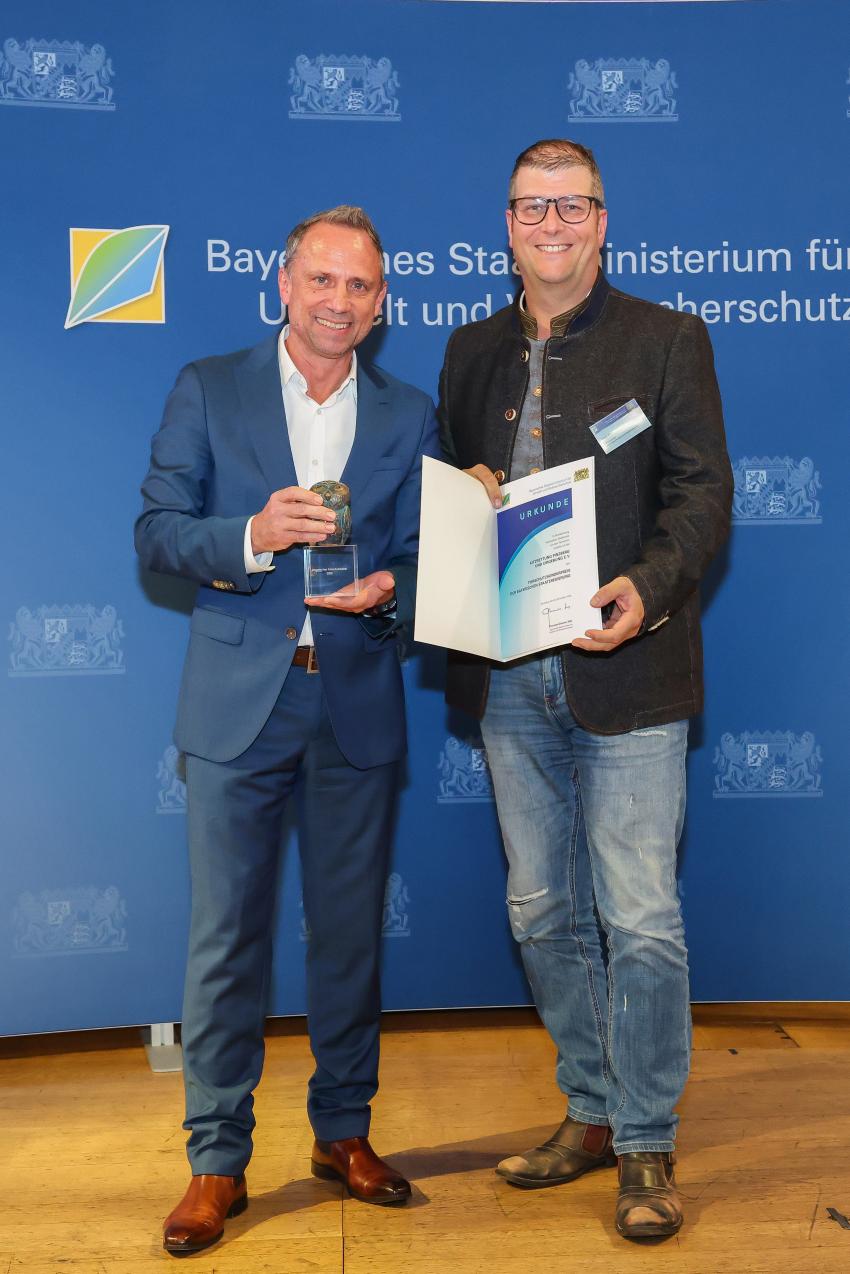 Das Foto zeigt den Bayerischen Umweltminister Thorsten Glauber mit einem Vertreter der Kitzrettung Pinzberg und Umgebung e. V., die Auszeichnungsurkunde und den Tierschutzpreis.