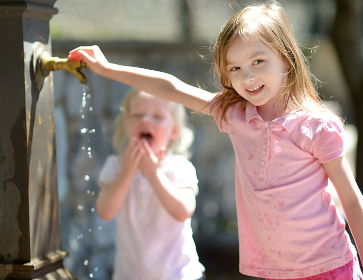 Das Bild zeigt zwei Mädchen an einem öffentlichen Trinkwasserbrunnen.