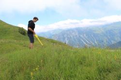 Mann steht auf einer Wiese mit einem langstieligem Gerät in der Hand vor Bergen