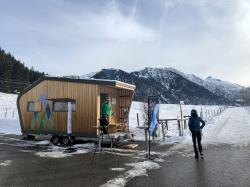 Eine kleine, modern gestaltete, mobile Holzhütte steht auf einem Parkplatz vor Bergpanorama