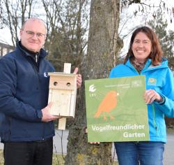 Ines Langensiepen steht rechts und hält das Modell einer Plakette vogelfreundlicher Garten in der Hand, Dr. Norbert Schäfer links mit einem Vogelhäuschen. Im Hintergrund sind Bäume erkennbar.