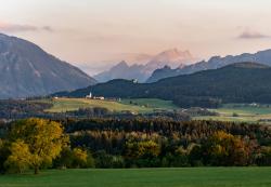 Landschaftsaufnahme bildet Wälder, Hügel, mittig ein Dorf in Miniaturansicht und im Hintergrund die Alpen ab.