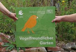 Zwei Hände halten eine quadratische Plakette, die ein stilisiertes Rotkehlchen zeigt und den Text Vogelfreundlicher Garten trägt.