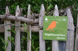Eine grüne Plakette mit Vogelgrafik und dem Schriftzug Vogelfreundlicher Garten hängt an einem Holzzaun.