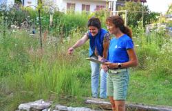  Zwei Frauen stehen in einem Garten und diskutieren, eine hält ein Klemmbrett in der Hand, die andere zeigt in den Garten.