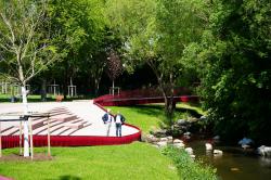 Zwei Männer stehen in einer Gartenanlage am Rande von amphietheaterartigen Stufen. Auf der anderen Seite schlängelt sich ein Bach entlang, frisch gepflanzte Bäume verweisen darauf, dass es sich um eine neue Anlage handelt, ein roter niedriger Zaun grenzt den Sitz- und Gehbereich vom Grün ab.
