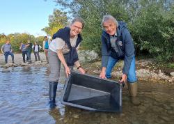 Eine Frau und ein Mann stehen mit Gummistiefeln im seichten Wasser und leeren einen großen Plastikbehälter aus.
