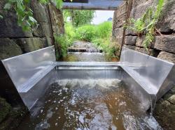  Eine U-förmige Metallstruktur im Wasser eines Wasserdurchlasses unter einer Brücke sorgt für eine Sperre im Bach.
