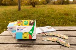 Zwei Spieleschachteln und das Begleitheft liegen auf einem Holztisch in einer Streuobstwiese.